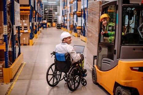 Großes Industriepalettenlager mit zwei Personen, die mit dem Gabelstapler und einem von ihnen im Rollstuhl arbeiten